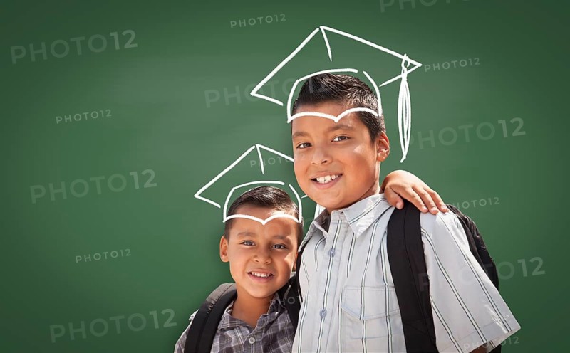 Young Hispanic Student Boys Wearing Backpacks In Front Of Blackboard with Graduation Caps Drawn In Chalk Over Head
