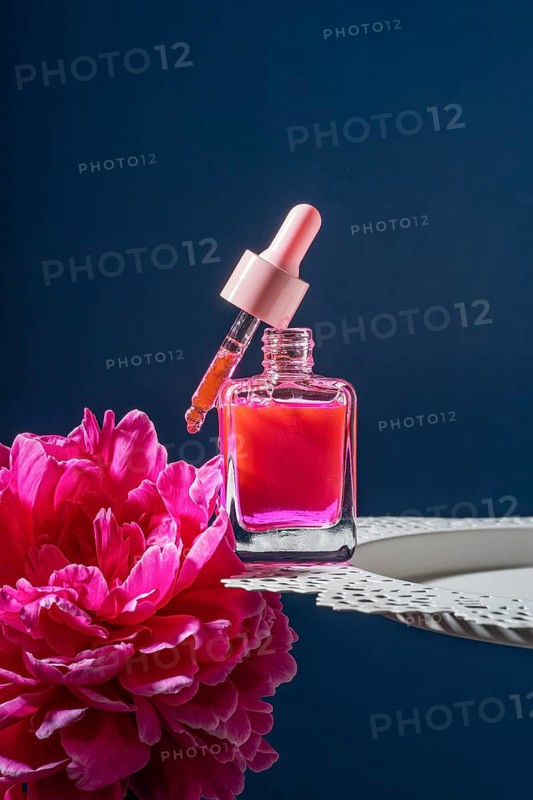 A vibrant pink serum in a transparent dropper bottle rests elegantly on a white plate, complemented by fresh pink peonies. The dark blue background enhances the scene's elegance