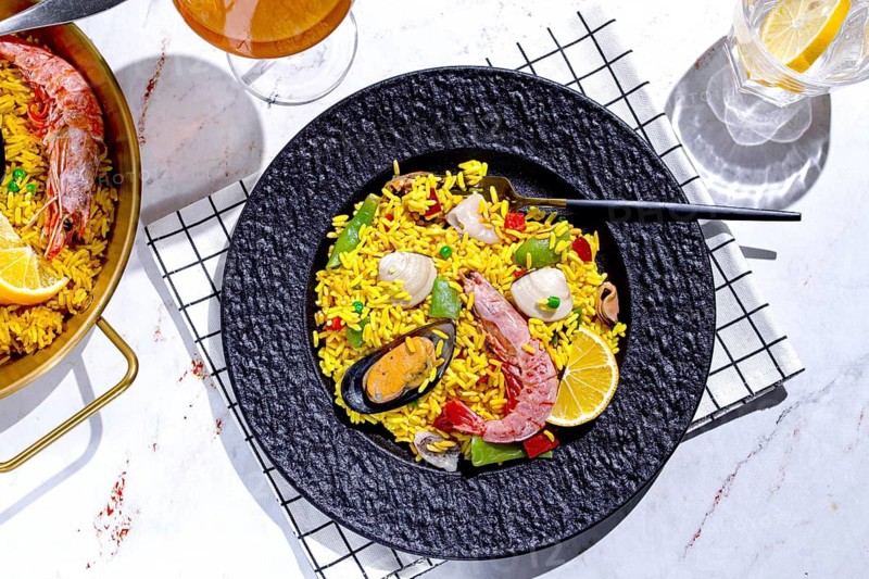 A table is set with a large black plate of seafood paella next to a golden pan. A glass with lemon slices sits nearby, along with another drink. Natural light fills the space