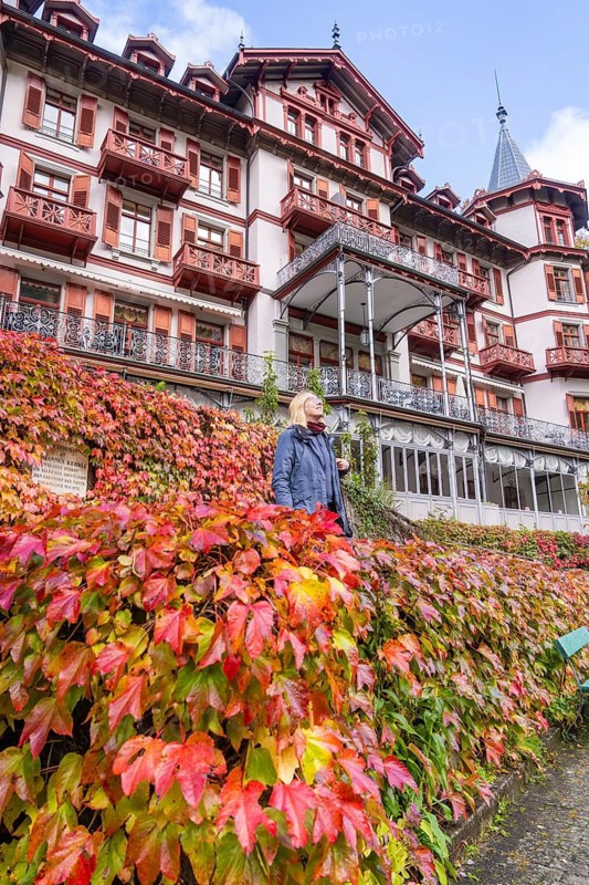 Woman in front of a hotel with a red facade, surrounded by autumn leaves and a bright sky, Grandhotel Giessbach, Lake Brienz, Switzerland