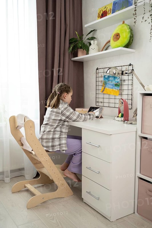 A child sits in a cozy room, using a tablet at a white desk. The room is decorated with playful stuffed toys and colorful artwork, creating a warm and inviting atmosphere