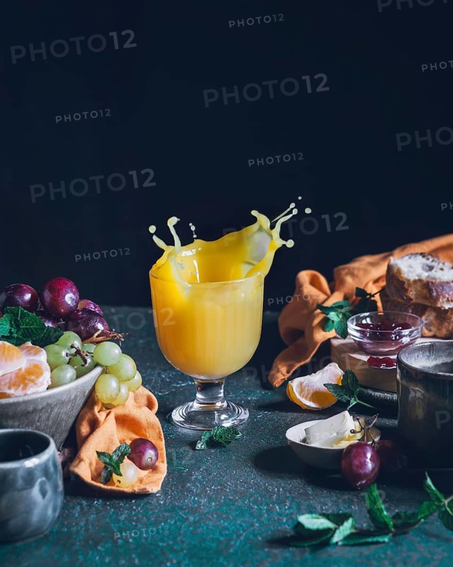 A vibrant splash of orange juice captured in mid-air in a glass goblet, surrounded by an assortment of fresh fruits, bread, and jars of preserves on a rustic table setting