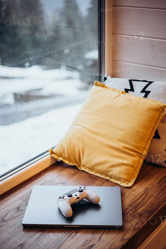 A cozy setup by a window with a gaming controller on a laptop, alongside a plush yellow pillow. Outside, a snowy landscape adds a serene, chill atmosphere