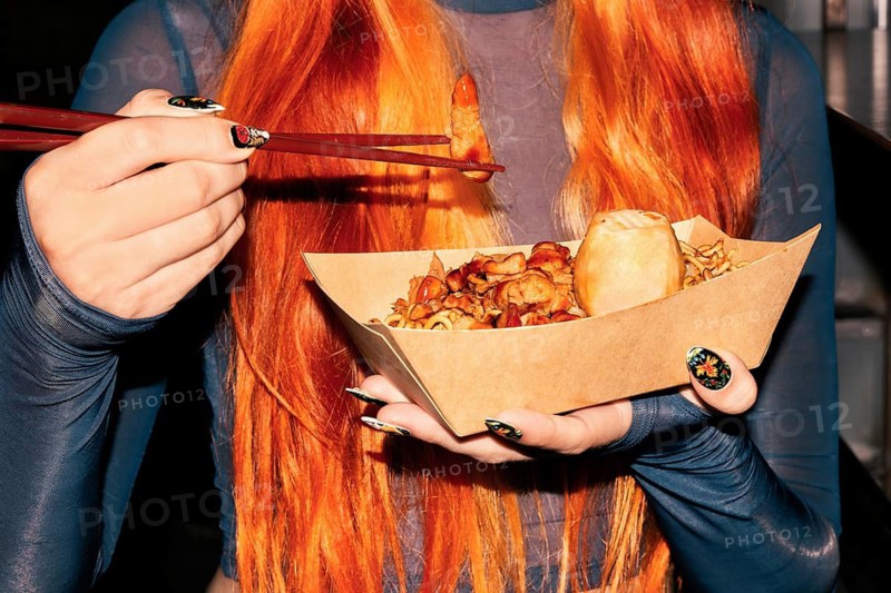 A woman with long orange hair is holding a paper tray of Asian street food, using chopsticks to eat She's at a vibrant food market, savoring delicious oriental flavors