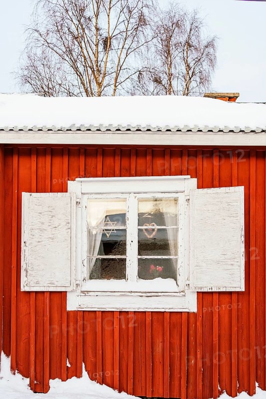 A picturesque view of a window on a red wooden cottage in Swedish Lapland, framed by frost and snow. Bare trees are visible in the background under a clear sky