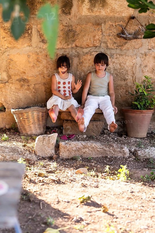 Two children sit on a stone bench against a rustic wall, surrounded by natural elements. They enjoy a sunny day together outdoors, creating a warm and joyful atmosphere