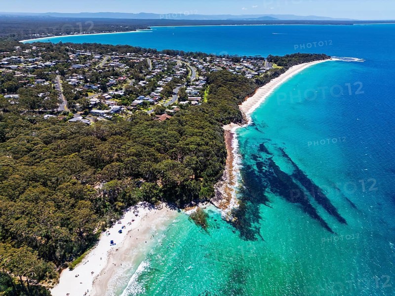 Aerial view of a coastal town featuring lush greenery, pristine beaches, and clear turquoise waters in Jervis Bay, Australia. Houses line the coast, offering a serene and picturesque landscape