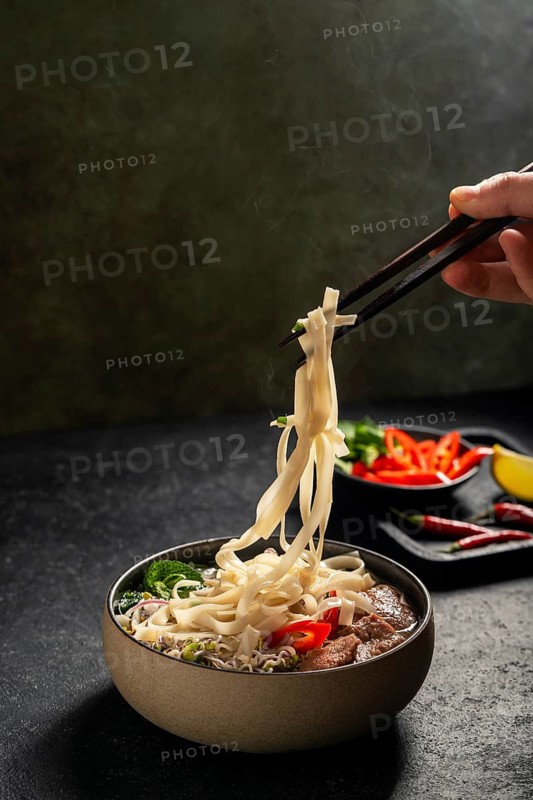 Cropped unrecognizable hand using chopsticks lifts steamy noodles from a bowl of Vietnamese beef pho, showcasing its rich broth and vibrant garnishes with chili peppers and herbs in the background