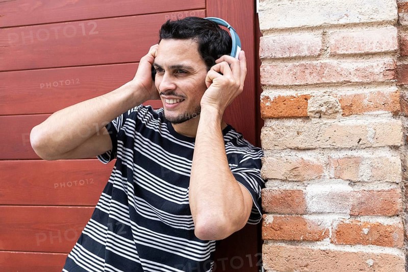 A cheerful cuban man wearing a striped shirt listens to music through headphones while leaning against a brick wall. His relaxed posture and happy expression convey a joyful and carefree mood