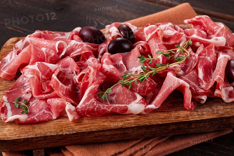 Freshly cut dried meat, served with olives and herbs, on a wooden chopping board, no people