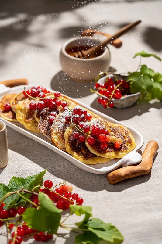 From above, a stack of golden pancakes is adorned with vibrant red currants and a sprinkle of powdered sugar, set on a white plate on a beige fabric background