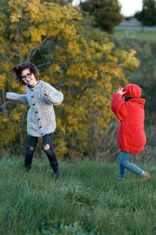 A girl and a boy enjoy a playful day in an autumnal park, surrounded by colorful foliage. The girl, in a red cardigan and glasses, smiles widely, while the boy in a grey sweater faces away