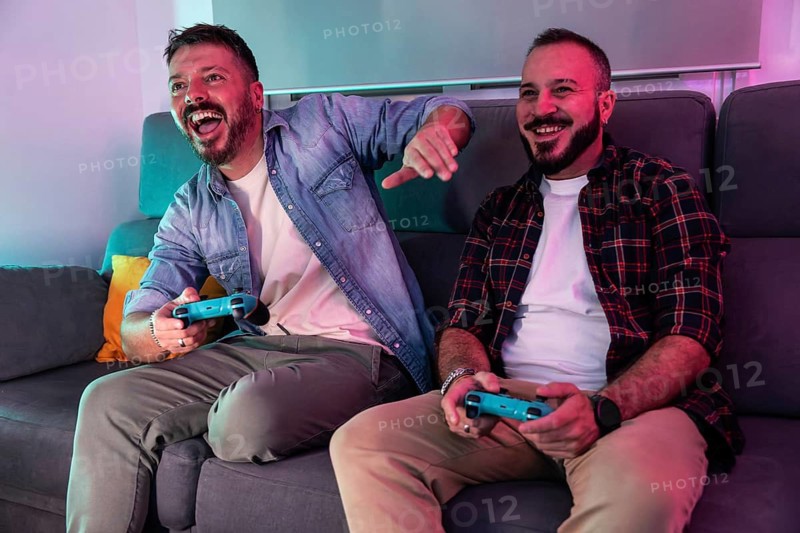 A joyful gay couple sitting on a couch, enthusiastically playing video games. They are relaxed and having fun, highlighting companionship and shared interests in a cozy home environment