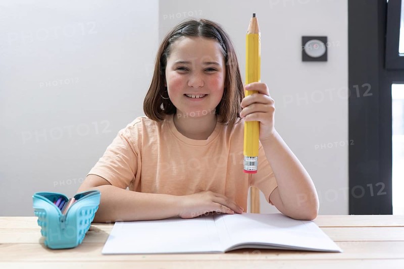 A smiling girl holds a giant pencil while sitting at a desk with an open notebook and a blue pencil case. The scene is bright and cheerful, capturing a study moment