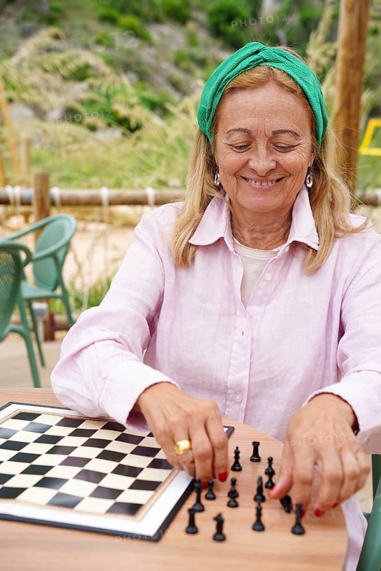 A woman sets up a chessboard on a wooden table in an outdoor setting, enjoying a leisurely game of chess Lush greenery and casual seating create a peaceful atmosphere