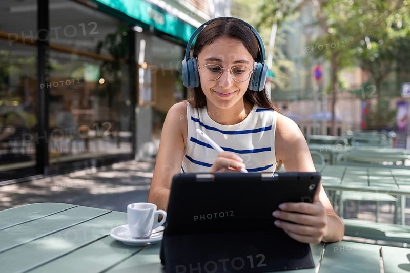 A woman wearing headphones sits at an outdoor cafe, using a digital tablet with a stylus. She enjoys a coffee, embodying the modern lifestyle in a vibrant city setting
