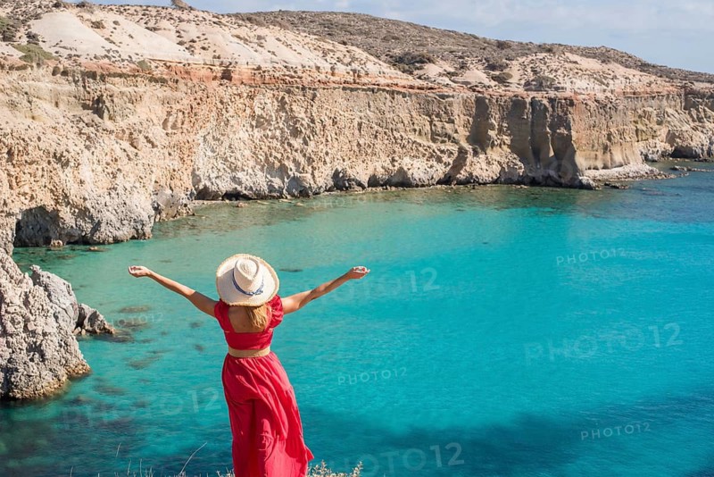 A woman in a red dress stands with arms outstretched, admiring the stunning turquoise waters and rugged cliffs of Tsigrado Beach on Milos Island, Greece