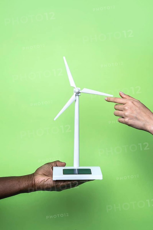 Close-up of two hands holding a small wind turbine model against a vibrant green background, symbolizing renewable energy and sustainability. The image captures innovation and eco-friendly solutions