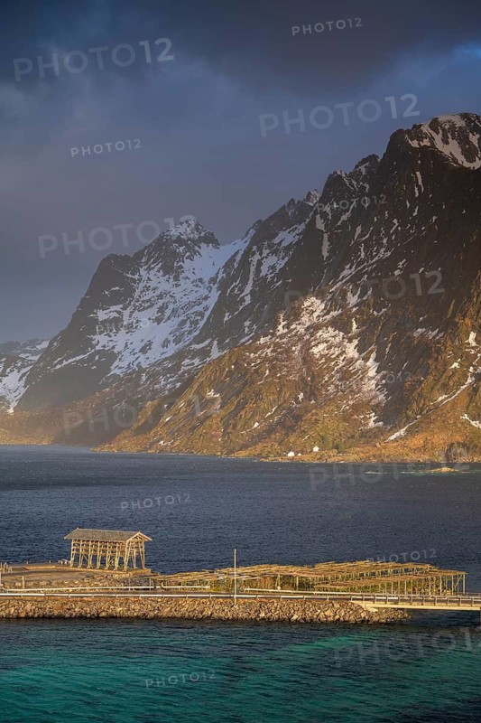 Racks for dried fish on Sakrisøy Island, snow-capped rocky mountains in the back, Sakrisoy, Reine, Lofoten, Norway