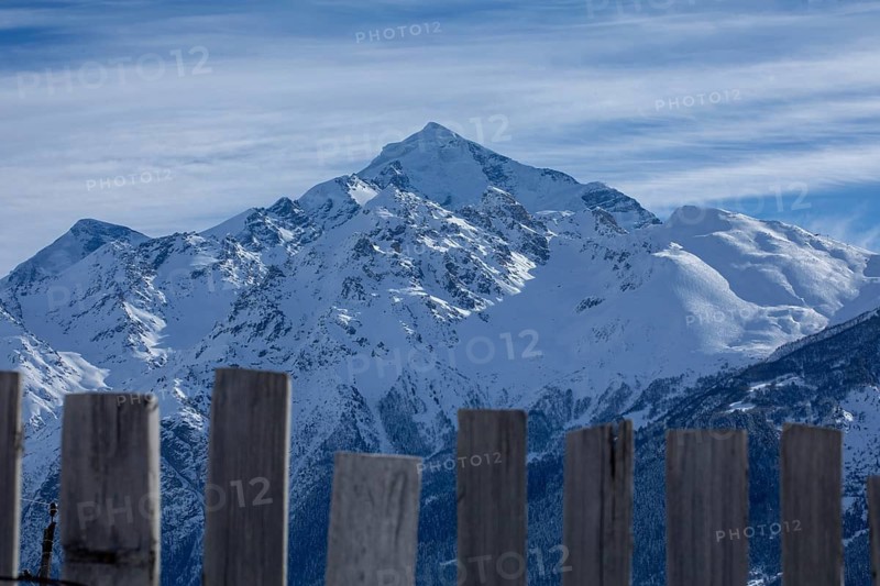 Majestic snowy peaks rise under a blue sky, framed by rustic wooden fence slats in the foreground, showcasing the serene beauty of a mountainous winter landscape in Georgia