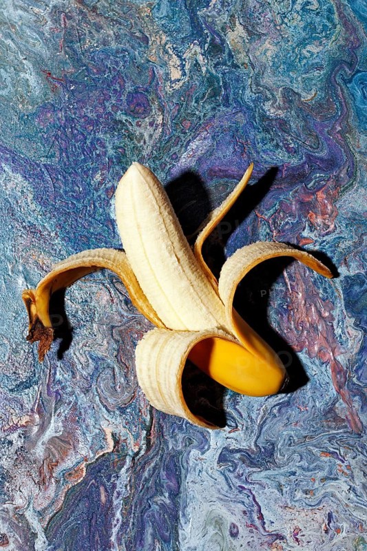 A peeled banana rests on a vibrant blue and purple swirled marble background. The contrast highlights the fruit fresh texture against the artistic design