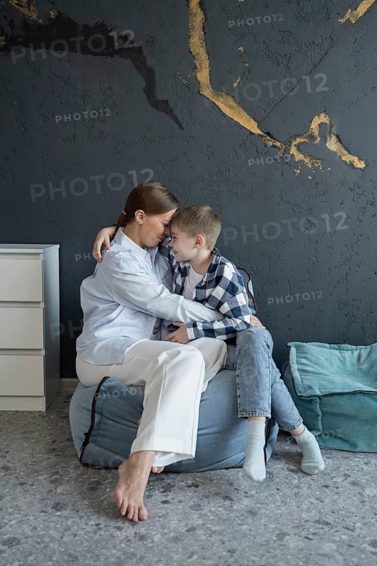 A mother and her young child share a heartfelt embrace, sitting together on cushions against a textured wall, expressing love and connection in a cozy, peaceful setting