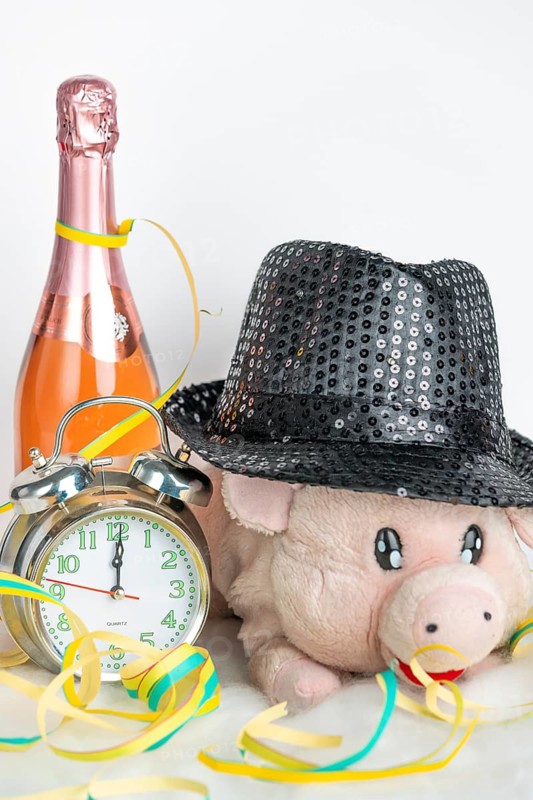 Plush pig wears glittering hat, accompanied by alarm clock and champagne bottle, New Year's Eve