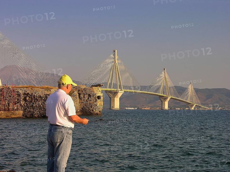 A man stands by the water and looks at a large suspension bridge, Rio-Antirrio cable car bridge, Rio fortress, Patras, Peloponnese, Greece