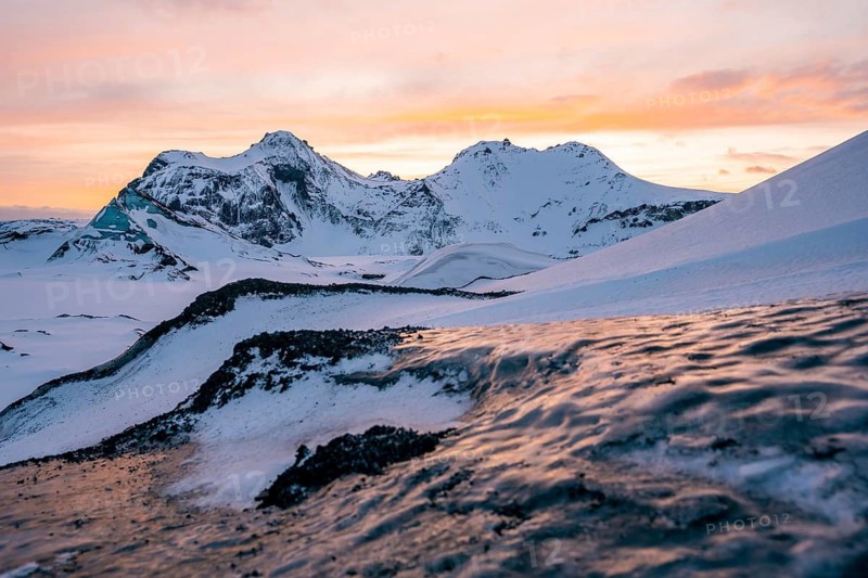 The snow-covered peaks of Katla Glacier glow under a soft orange-pink sunset, with textured volcanic ridges in the foreground creating dynamic contrast and depth in the wintry scene