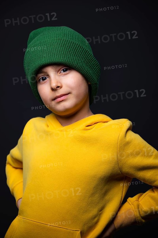 A child wearing a vibrant green beanie and a bright yellow hoodie confidently poses against a dark background. The image highlights a youthful and expressive personality