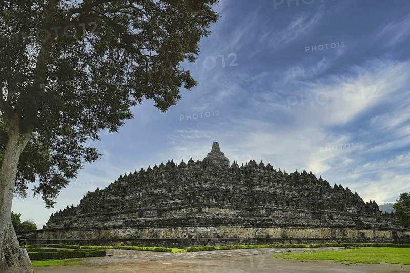 Majestic Borobudur Temple showcased under striking blue skies and wispy clouds, with lush greenery and ancient architectural details