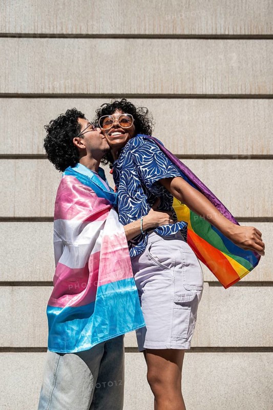 A multiethnic LGBTQ+ couple joyfully embracing, wrapped in vibrant Pride and transgender flags. Their cheerful expressions symbolize love, unity, and diversity