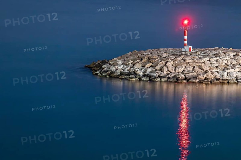 A serene lighthouse stands tall on a rocky shore at dusk, casting a warm glow across the calm Mediterranean Sea. The tranquil evening sky enhances the peaceful scenery