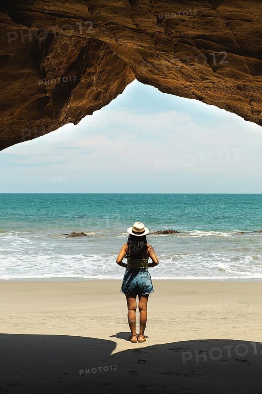 A young Latin woman stands on a picturesque beach in Mancora, Peru, enjoying the ocean view. She wears summer attire and a stylish hat, creating a serene coastal scene