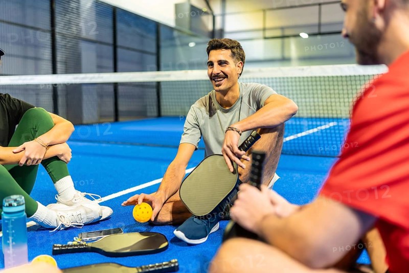 Diverse group of young adult friends enjoying a moment of rest and conversation on a blue pickleball court after playing, holding their paddles and ball