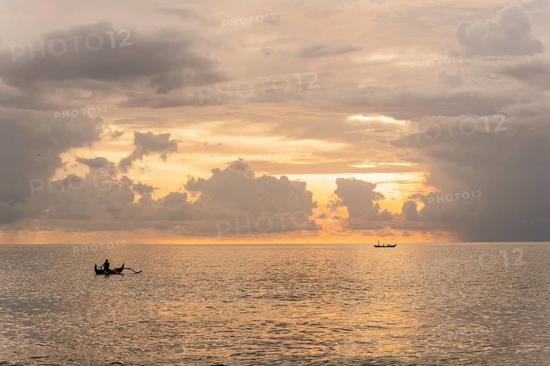 A serene sunset over the ocean in Bali, where fishermen navigate traditional boats on calm waters. The golden sky and scattered clouds create a peaceful and picturesque scene
