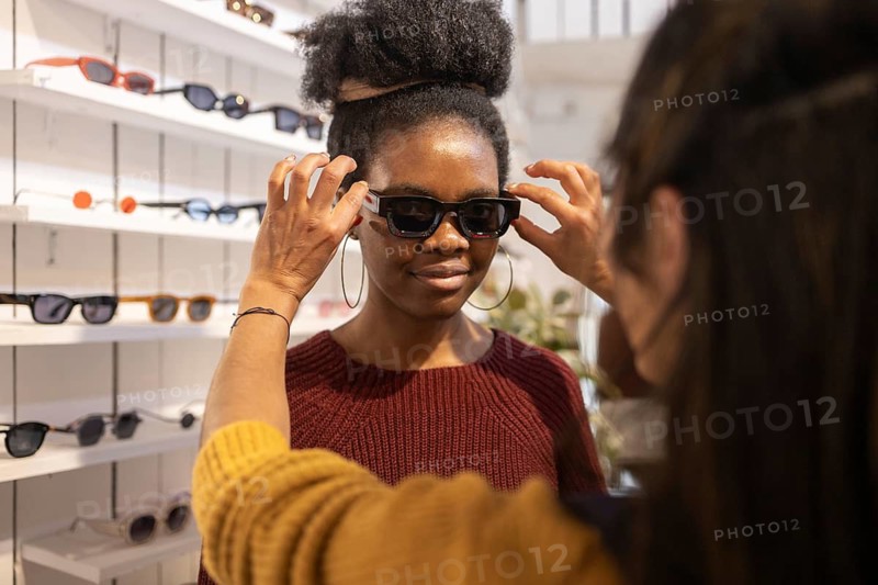 A woman tries on a pair of stylish sunglasses in a fashion boutique, assisted by an employee. The shelves are stocked with a variety of modern eyewear options, displaying a trendy selection