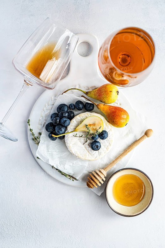 An elegant cheese platter featuring creamy brie cheese, fresh blueberries, and ripe pears, paired with honey and amber wine displayed on a white surface for a gourmet presentation
