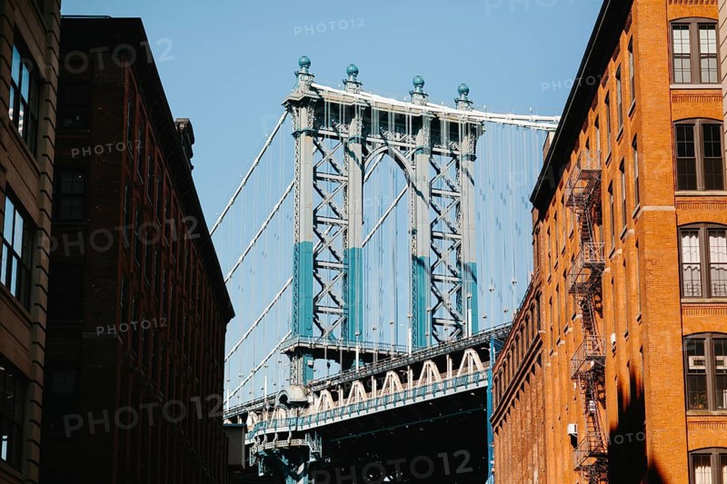 A striking view of a suspension bridge framed by two brick buildings in New York. The play of sunlight and shadows enhances the urban setting, emphasizing architectural details