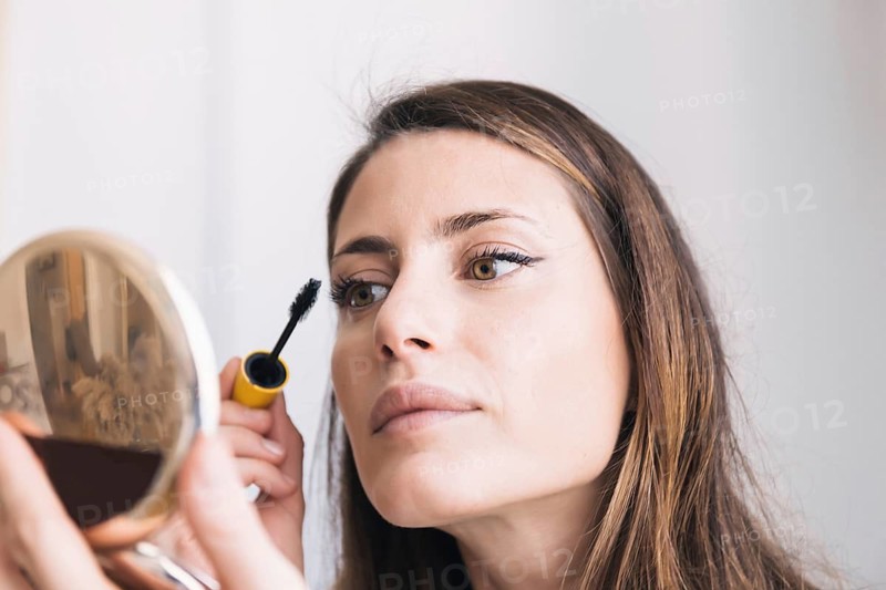 A woman applies mascara while looking into a mirror. Her face reflects concentration, highlighting the precision in her beauty routine, with a focus on her eyes and the makeup process