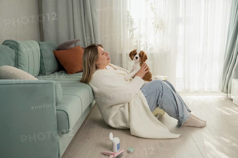A woman enjoys a cozy moment with her dog, sitting on the living room floor against a teal sofa. Sunlight filters through sheer curtains, highlighting the serene ambiance