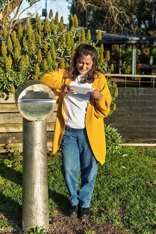A woman in casual attire stands by a modern mailbox in a lush Australian garden, enjoying her routine as she checks her mail amid greenery and sunlight