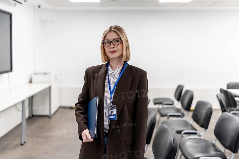 Young professional woman wearing glasses and a lanyard, holding a folder while standing confidently in a modern training room, ready for education or a business presentation