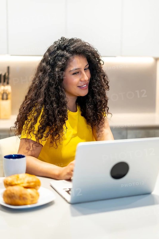 Vertical photo of a latin young woman eating breakfast and attending a video call sitting on the kitchen at home