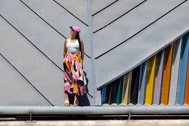 A woman stands confidently against a backdrop of modern architecture, her colorful outfit creating a striking urban contrast. She embodies a blend of style and structure