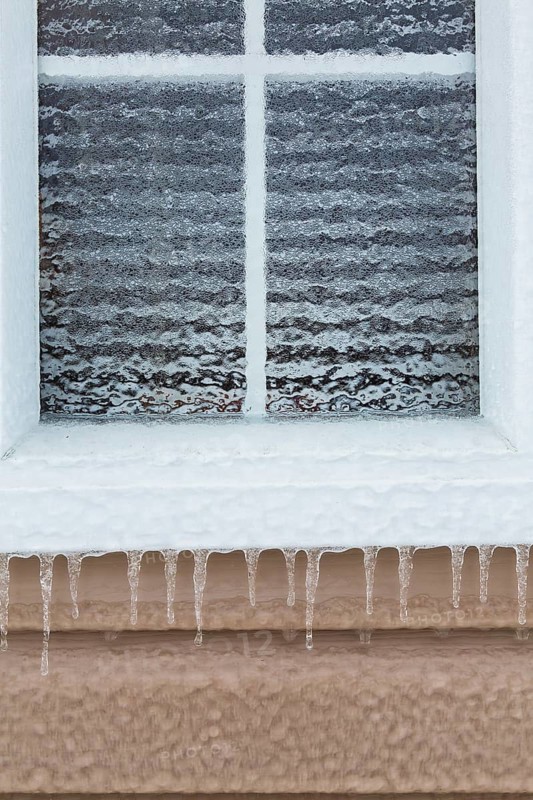 Close-up of glass pane window covered with thick sheet of ice and icicles hanging from edge of white frame after freezing rain in late winter, Quebec, Canada