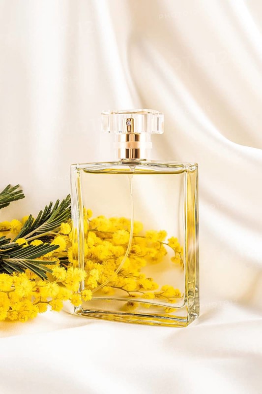 A sophisticated glass perfume bottle rests beside bright yellow mimosa flowers on elegant, creamy silk fabric, creating a serene and luxurious ambiance