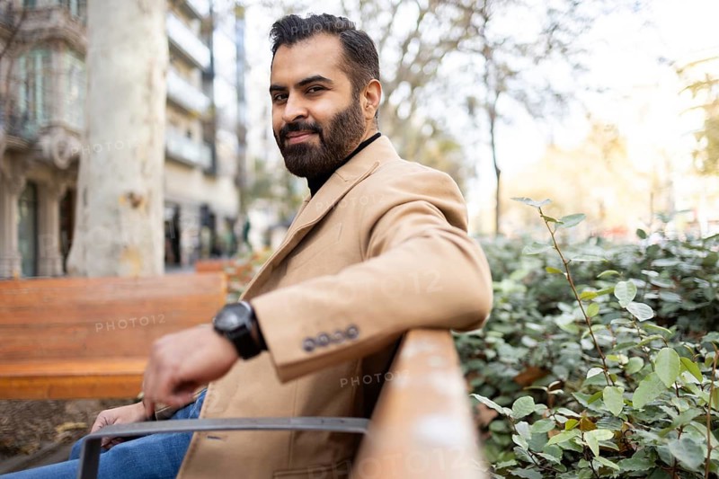 A cheerful Indian businessman in a stylish beige coat sits outdoors on a bench during a sunny winter day, looking at the camera with a friendly smile