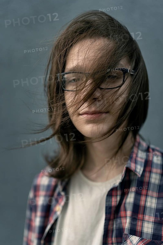 A teenage boy with long hair and glasses stands against a gray background, wearing a casual plaid shirt. His hair blows softly in the breeze, obscuring part of his face