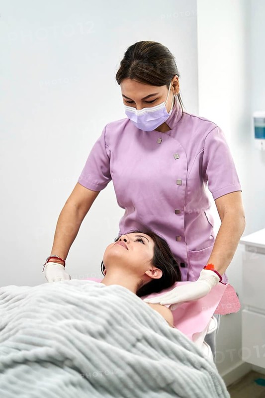 A beauty therapist in a lavender uniform and mask gently begins a facial treatment on a client, promoting relaxation and wellness in a clean, professional skincare clinic environment